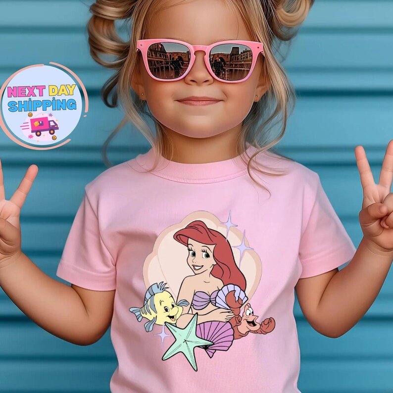 Puede incluir: Una camiseta rosa claro con una ilustraci&oacute;n de dibujos animados de Ariel de La Sirenita, con Flounder, Sebasti&aacute;n y una estrella de mar. La camiseta la lleva un ni&ntilde;o peque&ntilde;o con gafas de sol rosas.