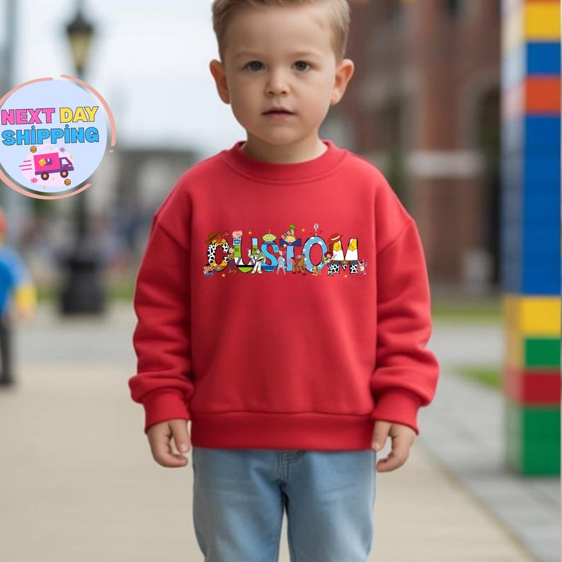 May include: A red sweatshirt with the word "CUSTOM" in colorful, cartoon-style letters. The sweatshirt is worn by a young child wearing blue jeans. The image includes a "NEXT DAY SHIPPING" logo in the upper left corner.