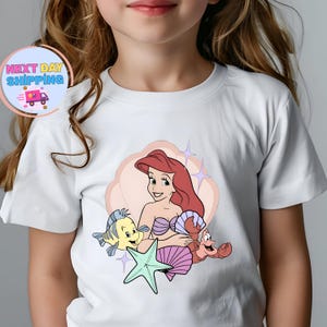 Puede incluir: Camiseta blanca con una ilustraci&oacute;n de dibujos animados de Ariel de La Sirenita, con Flounder, Sebasti&aacute;n y una estrella de mar. La imagen incluye el texto "Next Day Shipping" en un dise&ntilde;o circular con un cami&oacute;n de reparto.