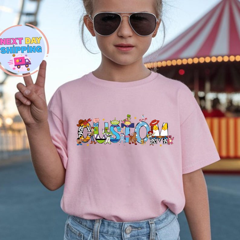 May include: A light pink t-shirt with the word "CUSTOM" in colorful letters. The letters are decorated with cartoon characters. The shirt is worn by a person wearing sunglasses.