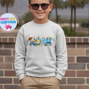 May include: A light gray sweatshirt with the name "NOAH" in colorful, cartoon-style letters. The letters are decorated with characters from a popular animated movie. The sweatshirt is worn by a person.