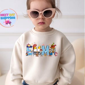 May include: A cream-colored sweatshirt featuring the name "EMMA" in blue letters with Toy Story character designs. The sweatshirt is worn by a young child wearing pink sunglasses.