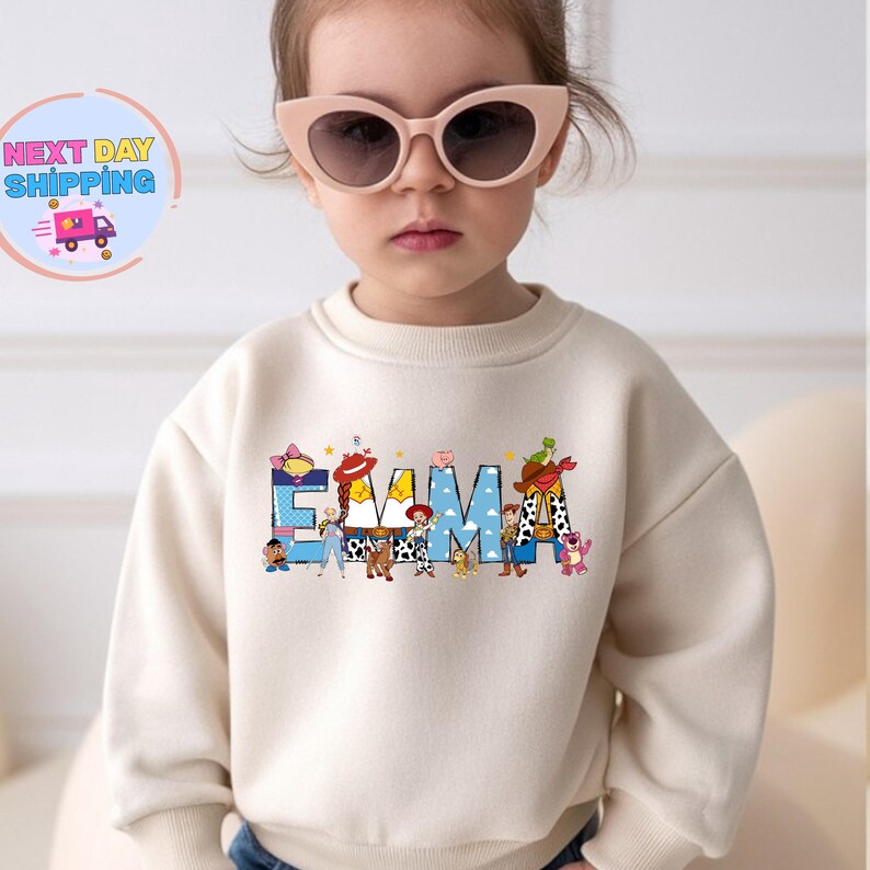 May include: A cream-colored sweatshirt featuring the name "EMMA" in blue letters with Toy Story character designs. The sweatshirt is worn by a young child wearing pink sunglasses.