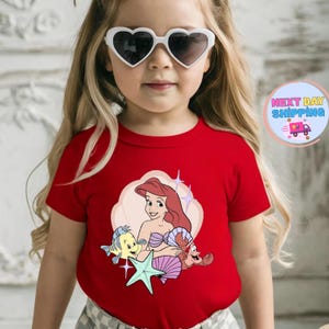 Puede incluir: Una camiseta roja con una ilustraci&oacute;n de dibujos animados de Ariel de La Sirenita, con Flounder, Sebasti&aacute;n y una estrella de mar. El dise&ntilde;o est&aacute; sobre un fondo rosa claro en forma de nube. La camiseta la lleva un ni&ntilde;o con gafas de sol en forma de coraz&oacute;n.