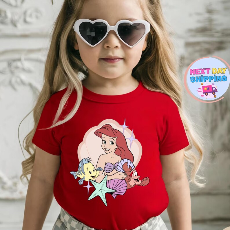 Puede incluir: Una camiseta roja con una ilustraci&oacute;n de dibujos animados de Ariel de La Sirenita, con Flounder, Sebasti&aacute;n y una estrella de mar. El dise&ntilde;o est&aacute; sobre un fondo rosa claro en forma de nube. La camiseta la lleva un ni&ntilde;o con gafas de sol en forma de coraz&oacute;n.