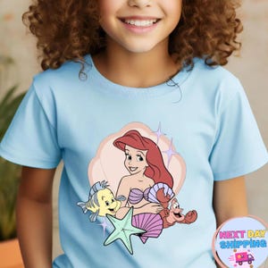 Puede incluir: Camiseta azul claro con un dise&ntilde;o de Ariel de La Sirenita. El dise&ntilde;o incluye a Ariel, Flounder, Sebasti&aacute;n y una estrella de mar. La camiseta tiene cuello redondo y mangas cortas.