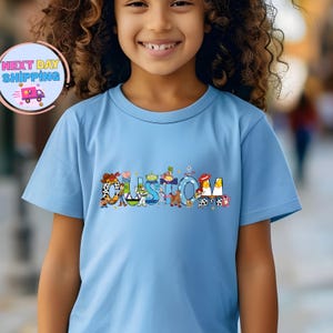 May include: A light blue t-shirt with the word "CUSTOM" in colorful letters, each designed with characters from a popular animated movie. The shirt is worn by a young person.