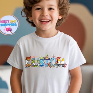 May include: White t-shirt with the word "CUSTOM" in colorful, cartoon-style letters. The letters are decorated with characters from a popular animated movie. The shirt is worn by a smiling child.