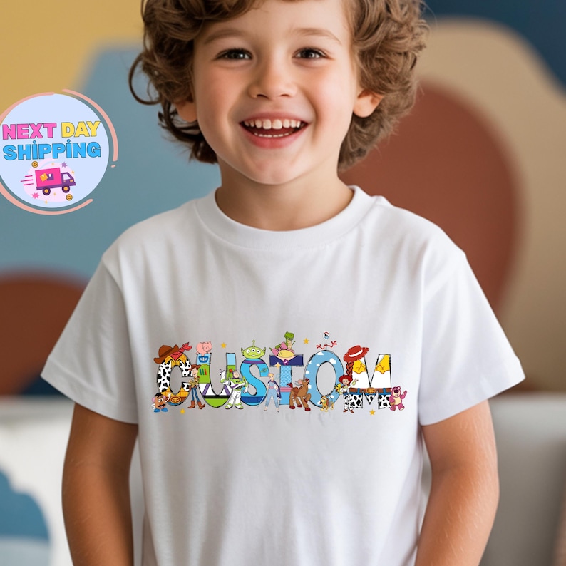 May include: White t-shirt with the word "CUSTOM" in colorful, cartoon-style letters. The letters are decorated with characters from a popular animated movie. The shirt is worn by a smiling child.