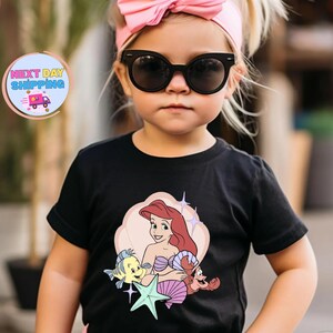 Puede incluir: Una camiseta negra con un gr&aacute;fico colorido de Ariel de La Sirenita, con Flounder y Sebasti&aacute;n. El dise&ntilde;o est&aacute; sobre un fondo de color melocot&oacute;n claro. El ni&ntilde;o lleva una diadema rosa y gafas de sol.
