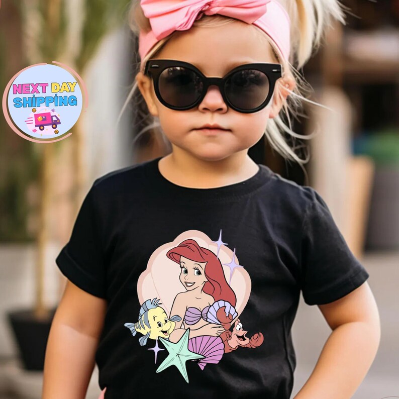 Puede incluir: Una camiseta negra con un gr&aacute;fico colorido de Ariel de La Sirenita, con Flounder y Sebasti&aacute;n. El dise&ntilde;o est&aacute; sobre un fondo de color melocot&oacute;n claro. El ni&ntilde;o lleva una diadema rosa y gafas de sol.