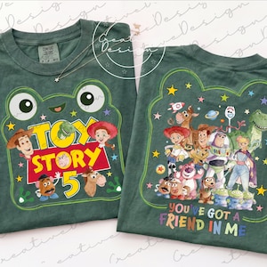 May include: Two dark green t-shirts featuring Toy Story graphics. The left shirt displays a frog-shaped frame with the text Toy Story 5 and character faces. The right shirt shows a group of Toy Story characters and the text You've Got A Friend In Me.