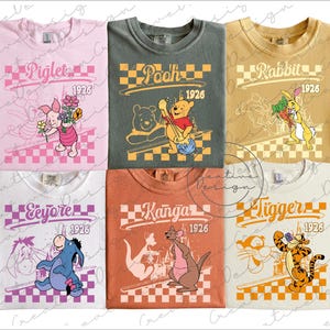 May include: Six colorful t-shirts featuring Winnie the Pooh characters. Each shirt displays a character's name and "1926" in a checkered design. Characters include Piglet, Pooh, Rabbit, Eeyore, Kanga, and Tigger. The shirts are in various colors.