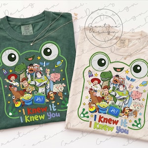 以下が含まれることがあります： ダークグリーンとクリーム色のクルーネックTシャツ2枚。どちらもトイ・ストーリーのキャラクターが描かれたカエルの顔の大きなグラフィックが特徴です。カエルの下には「I Knew It I Knew You」というテキストがカラフルな文字でプリントされています。