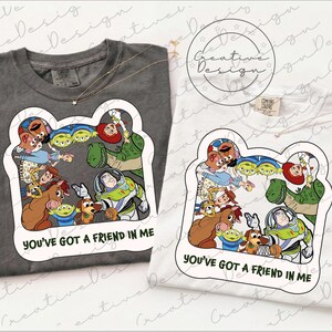 May include: A charcoal grey t-shirt and a white t-shirt, each featuring a graphic print of various animated characters from Toy Story arranged in a circle, with the text YOU'VE GOT A FRIEND IN ME printed in black below the character illustration.
