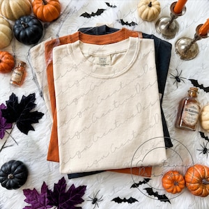 Könnte beinhalten: Ein Stapel aus drei gefalteten T-Shirts in den Farben Creme, Orange und Schwarz. Das oberste cremefarbene T-Shirt hat ein Comfort Colors Etikett. Die Shirts sind umgeben von kleinen Kürbissen, zwei Apothekerflaschen mit den Etiketten Arsenic und Belladonna sowie Fledermäusen.