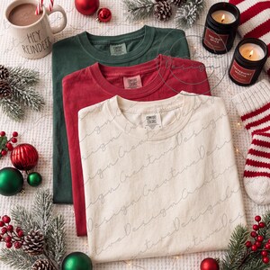 Könnte beinhalten: Drei T-Shirts mit Rundhalsausschnitt in den Farben Creme, Rot und Dunkelgrün, jeweils mit einem Etikett der Marke Comfort Colors am inneren Halsausschnitt. Die T-Shirts sind in einem gestapelten, überlappenden Layout auf einer strukturierten weißen Oberfläche angeordnet.