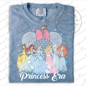 May include: A light blue t-shirt featuring Disney princesses with a pink bow, two white sphere shapes, and the text "Princess Era." A gold necklace with a pendant is on top of the shirt.