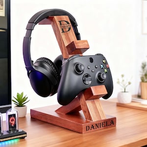 May include: A wooden headphone and game controller stand with the name "Daniela" engraved on the base. The stand holds a black headset and a black game controller. The wood has a reddish-brown finish. The name "Daniela" is also engraved on the top of the stand.