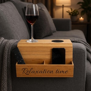 Puede incluir: Una mesa de reposabrazos de sofá de bambú con una copa de vino, un mando a distancia y un smartphone. La mesa tiene un portavasos y un compartimento con las palabras "Relaxation time" grabadas. La mesa está colocada sobre un sofá gris.