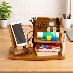 May include: A wooden desk tidy with various compartments. It includes a phone stand, a section for glasses, and a space for watches. The organiser also has a small tray with colourful items and a book. The words "BEST DAD EVER" are engraved on the back.