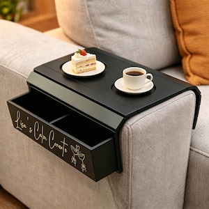 Puede incluir: Bandeja negra para reposabrazos de sofá con cajón incorporado. La bandeja contiene un plato blanco con una porción de pastel y una taza de café. El cajón tiene el texto "Lisa's Calm Corner" en letras doradas. La bandeja descansa sobre un sofá gris.