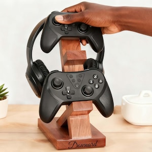 Op de afbeelding: Een houten standaard voor gamecontrollers en een koptelefoon, met een donkere afwerking. Twee zwarte controllers en een zwarte koptelefoon worden getoond. De standaard heeft de inscriptie "Downard".