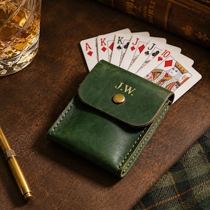 May include: A dark green leather card holder with a flap closure secured by a brass-colored snap button. The flap is embossed with the initials J.W. in gold lettering. A hand of playing cards is tucked behind the wallet, showing a royal flush.