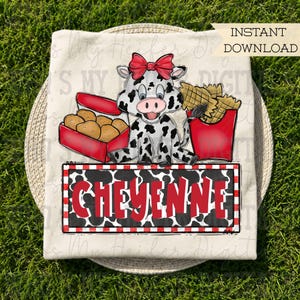 May include: A cream-colored t-shirt featuring a graphic of a spotted cow wearing a red bow, sitting between a red box of chicken nuggets and a red container of french fries. Below the cow is a rectangular sign with the name CHEYENNE in red.
