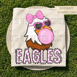 May include: A cream-colored t-shirt featuring a graphic of a bald eagle wearing a pink bow and heart-shaped sunglasses while blowing a pink bubble. Below the eagle, the word EAGLES is printed in a pink and white checkered font pattern.