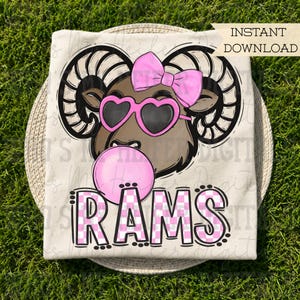 Könnte beinhalten: Ein beiges T-Shirt mit einer Grafik eines braunen Widders mit großen, geschwungenen Hörnern, der eine rosa Schleife und eine rosa herzförmige Sonnenbrille trägt und eine rosa Kaugummiblase bläst. Der Text RAMS steht darunter in rosa-weißem Karomuster.