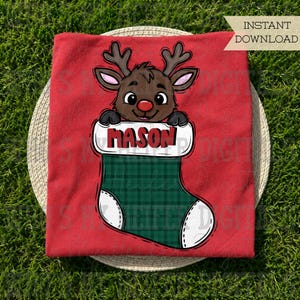 May include: A red t-shirt featuring a graphic of a brown reindeer with antlers peeking out of a green plaid Christmas stocking. The stocking has a white cuff with the name MASON printed in bold red letters. The design includes a red nose.