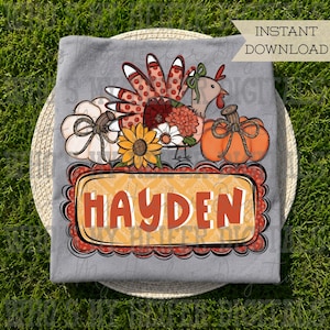 May include: A gray t-shirt featuring a graphic of a turkey with floral feathers, a white pumpkin, an orange pumpkin, and a sunflower. Below the graphic is a rectangular frame with a scalloped border containing the orange text HAYDEN.