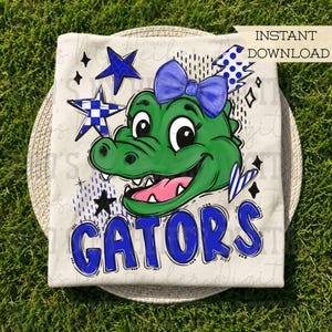 Peut inclure: Un t-shirt couleur crème avec un motif d'alligator vert portant un nœud bleu, entouré d'étoiles bleues et de motifs à carreaux. Le mot GATORS est imprimé en grandes lettres bleues en bas. Une bannière INSTANT DOWNLOAD est en haut à droite.