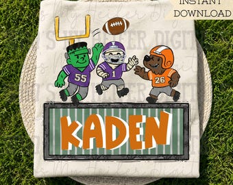 May include: A cream-colored t-shirt featuring a graphic of a Frankenstein monster, a mummy, and a bear playing football. The characters wear jerseys numbered 55, 1, and 26. Below them is a green field graphic with the name KADEN in orange letters.