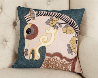May include: Decorative square pillow with a teal background and an embroidered horse head design. The horse is cream-colored with brown, yellow, and black accents, and floral details. The pillow is placed on a beige upholstered chair.