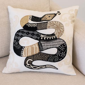 May include: A decorative square pillow with a detailed embroidered snake design. The snake features black, tan, and cream-colored patterns. The pillow is on a cream-colored sofa, and the background has a subtle floral pattern.