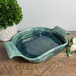 May include: A rectangular ceramic baking dish with integrated side handles, featuring a glossy, deep forest green glaze with lighter teal and blue-green mottled variations across the surface and edges, showcasing a smooth, reflective finish.