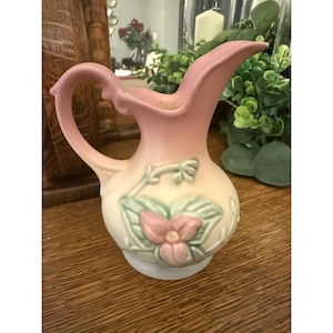 May include: A vintage pink and cream-colored ceramic pitcher. The pitcher features a floral design with a pink flower and green leaves. It has a curved handle and a spout for pouring. The pitcher is likely used for serving beverages.