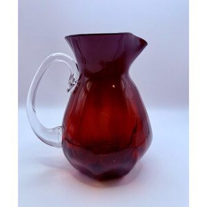 May include: A ruby red glass pitcher with a clear glass handle. The pitcher has a rounded body and a flared spout. The handle is attached to the body with a small decorative detail. The pitcher is likely used for serving beverages.