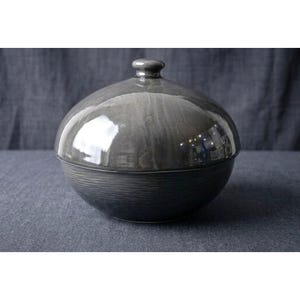 May include: A round ceramic lidded bowl with a glossy grey finish. The lid is smooth and dome-shaped with a small knob handle at the top. The base of the bowl features a textured, horizontal ribbed pattern in a darker shade of charcoal grey.
