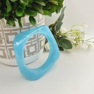 May include: A light blue, square-shaped bangle bracelet with a smooth, glossy finish and subtle marbled texture. The bracelet features a thick, rounded profile and a translucent appearance, showcasing a consistent sky blue color throughout.