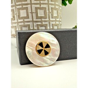 May include: A round, iridescent white brooch with a gold-tone center. The center features a black and crystal design. The brooch is set on a black box, with a white background and a decorative vase in the background.