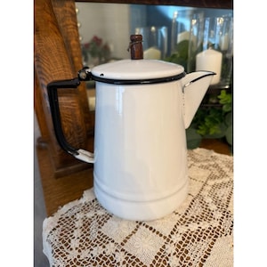 May include: A white enamel coffee pot with a black handle and trim. The pot has a wooden knob on the lid and a spout for pouring. The pot is sitting on a white lace tablecloth.