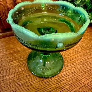 May include: A green glass pedestal bowl with a scalloped rim and decorative leaf pattern. The bowl has a round base and a central pedestal, showcasing a vintage aesthetic. The bowl is approximately 6 inches in diameter.