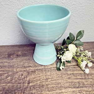 May include: A light blue ceramic bowl with a pedestal base. The bowl has a wide opening and a rounded shape. The surface has a textured, hand-painted appearance. White flowers and greenery are arranged beside the bowl on a wooden surface.