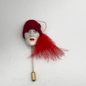 May include: A lapel pin featuring a sculpted face wearing a deep red felt cloche hat and a black net veil. The face has painted features including red lips and dark eyes. A cluster of bright red feathers extends from the side of the hat.