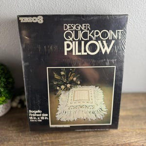 May include: A black rectangular box for a Trio 3 Designer Quickpoint Pillow kit. The packaging features white text and a photograph of a finished off-white square pillow with fringe and geometric patterns, noting a finished size of 18 in. x 18 in.