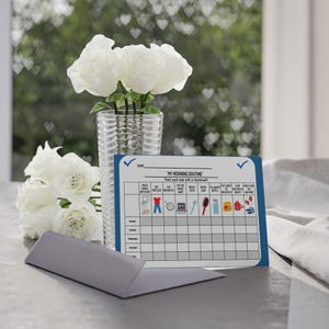 Könnte beinhalten: Eine Morgenroutine-Tabelle mit blauem Rand und dem Text "MY MORNING ROUTINE". Die Tabelle enthält illustrierte Aufgaben wie Händewaschen, Anziehen und Zähneputzen. Eine Vase mit weißen Rosen steht in der Nähe.