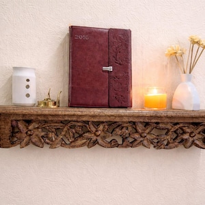 Peut inclure: Une étagère en bois sculpté de motifs floraux supporte un journal en cuir bordeaux, un récipient en céramique blanc, une bougie allumée et un petit vase avec des fleurs séchées. L'étagère est fixée à un mur crème.